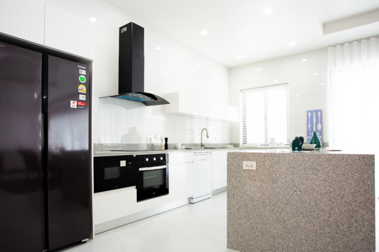A modern kitchen with white cabinets, built-in oven, stovetop with range hood, refrigerator, granite island counter, and bright natural lighting.