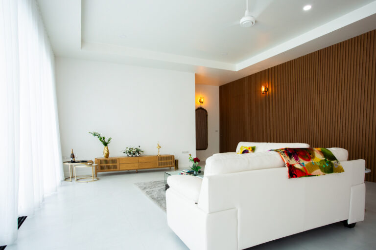 A bright modern living room with a white sofa, wooden accent wall, minimal decor, and large windows with sheer curtains allowing natural light inside.