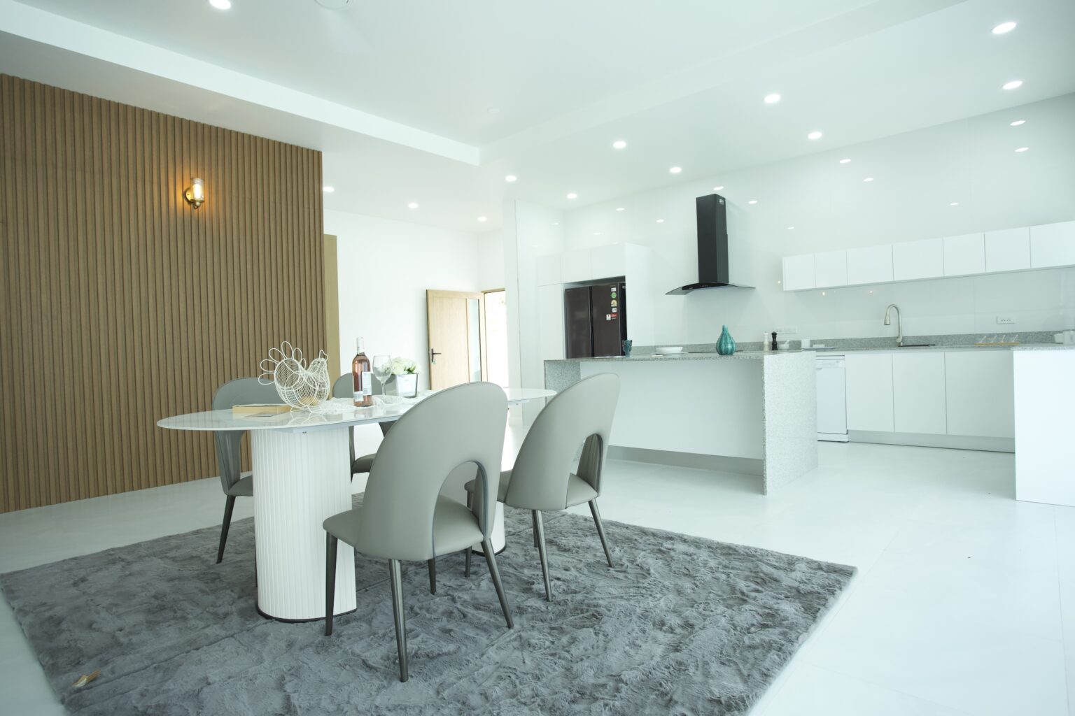 Modern open-concept dining area with a white oval table, grey designer chairs on a plush rug, and a sleek white minimalist kitchen at Villa de l'Amitie in Hua Hin.