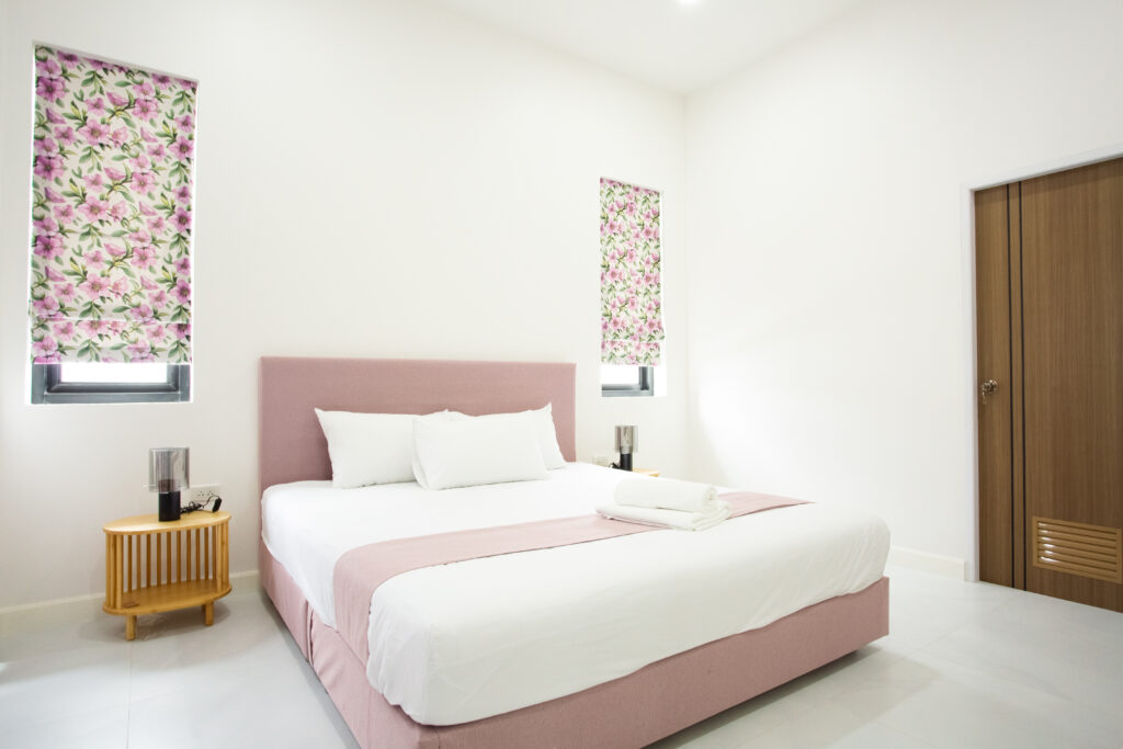A modern bedroom with a pink upholstered bed, white bedding, floral window curtains, a wooden bedside table, and a wooden door in a clean, minimal interior.