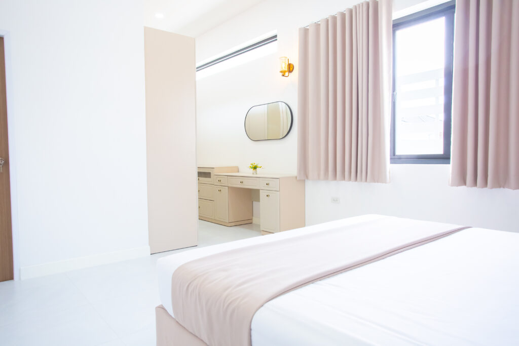 A wide-angle, bright photo of a minimalist style Master Bedroom with light wood furniture, a built-in dressing table with an oval mirror, light curtains, and a partially seen large bed, showcasing ample space.