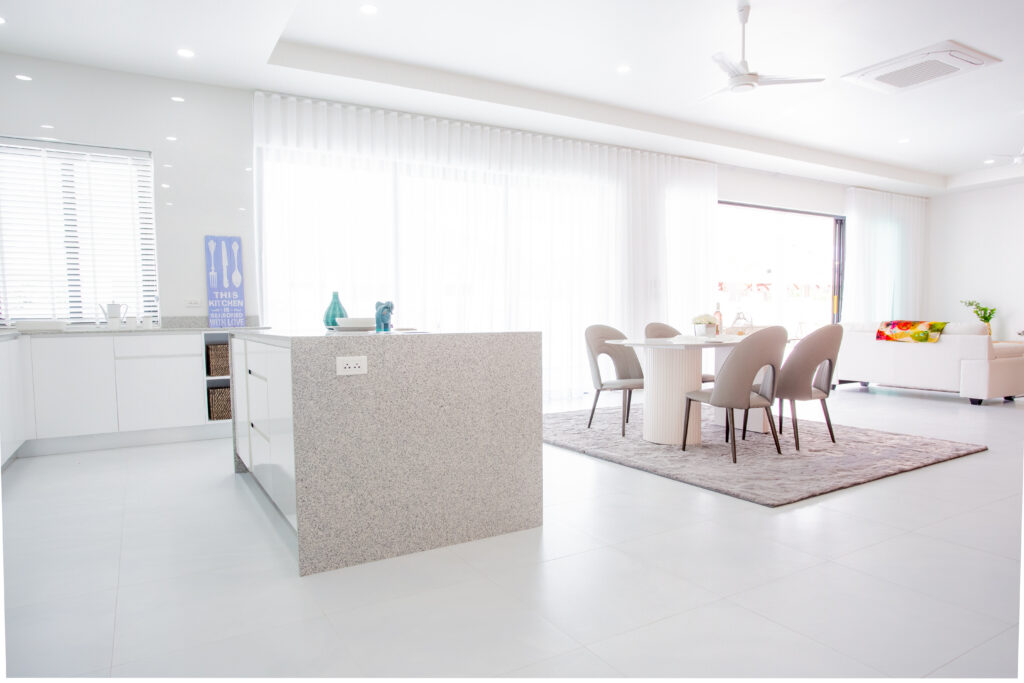 A bright and spacious open-plan interior featuring a granite kitchen island, a modern dining set on a plush rug, and large floor-to-ceiling glass doors with sheer curtains at Villa de l'Amitie in Hua Hin.