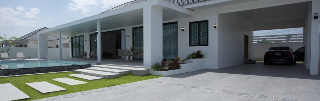 Wide-angle view of a modern white pool villa featuring a private swimming pool, sun loungers, an attached carport with a car, and a landscaped garden under a cloudy blue sky at Villa de l'Amitie in Hua Hin.