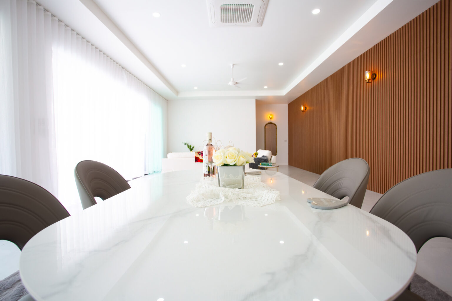A modern dining area with a long white marble table, cushioned chairs, decorative centerpiece, wooden accent wall, and large windows with sheer curtains allowing natural light.