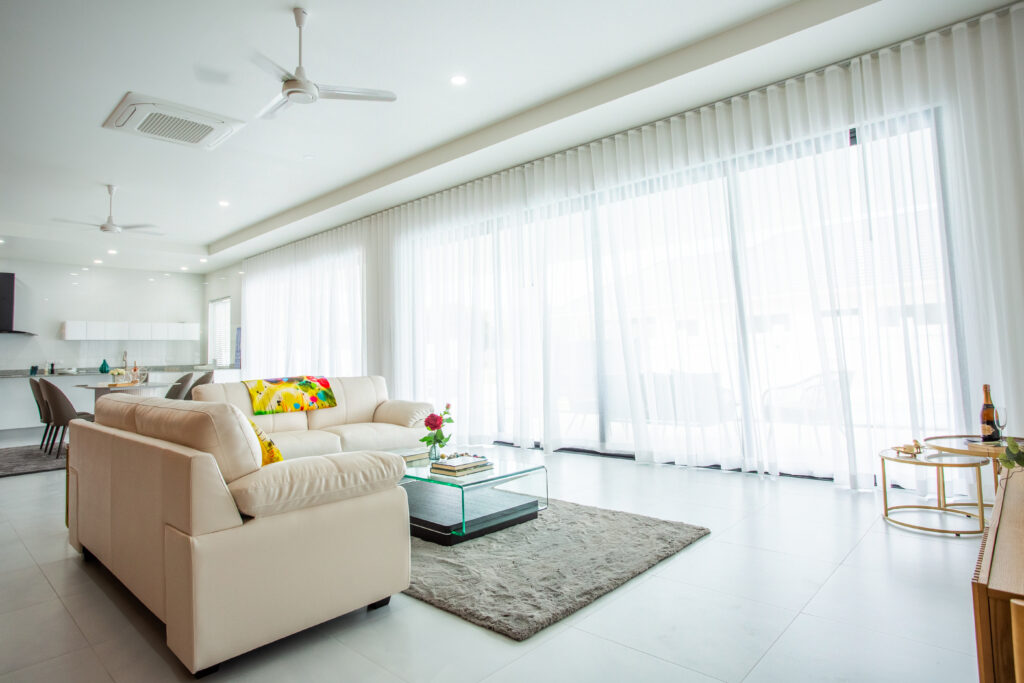 Bright living room with beige sofa, glass coffee table, and floor-to-ceiling sheer curtains allowing natural light to fill the space.