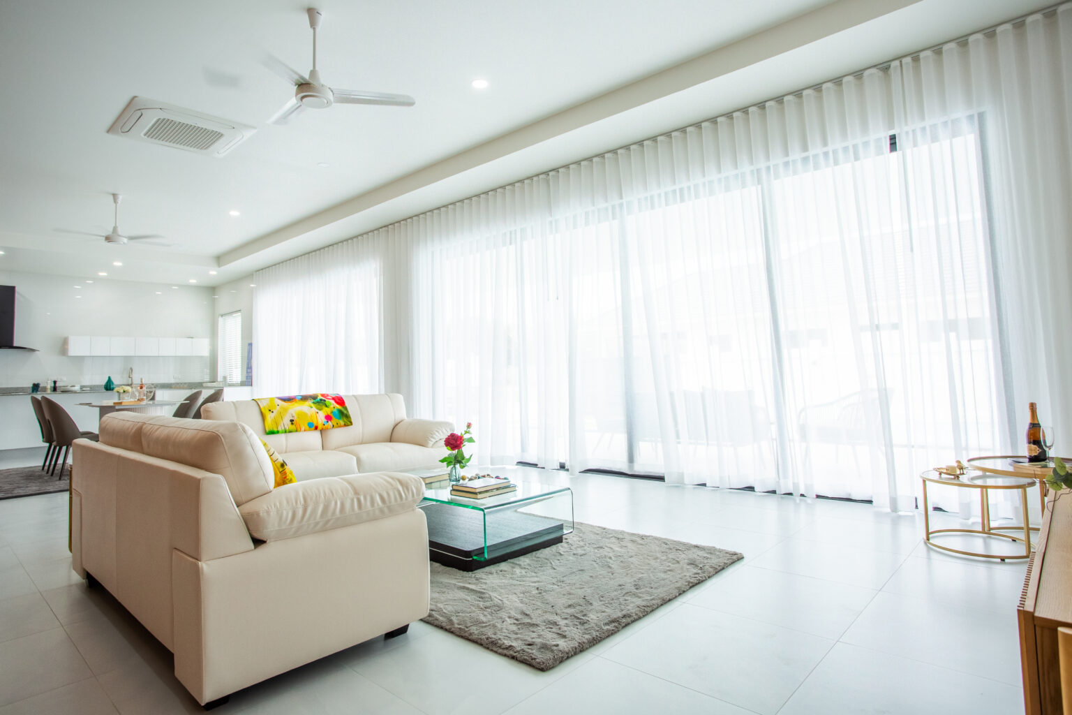 Bright living room with beige sofa, glass coffee table, and floor-to-ceiling sheer curtains allowing natural light to fill the space.