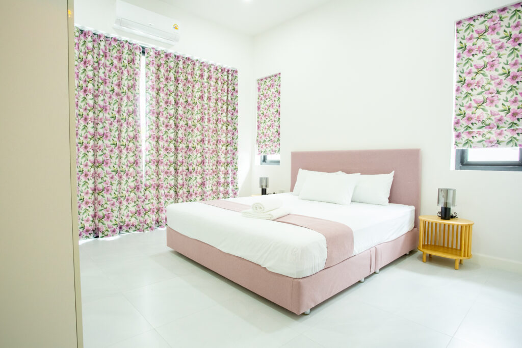 A bright and airy bedroom featuring a pastel pink bed, floral curtains, and white walls at Villa de L’Amitié Huahin.