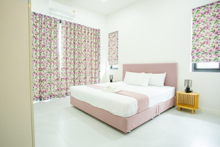 A bright and airy bedroom featuring a pastel pink bed, floral curtains, and white walls at Villa de L’Amitié Huahin.