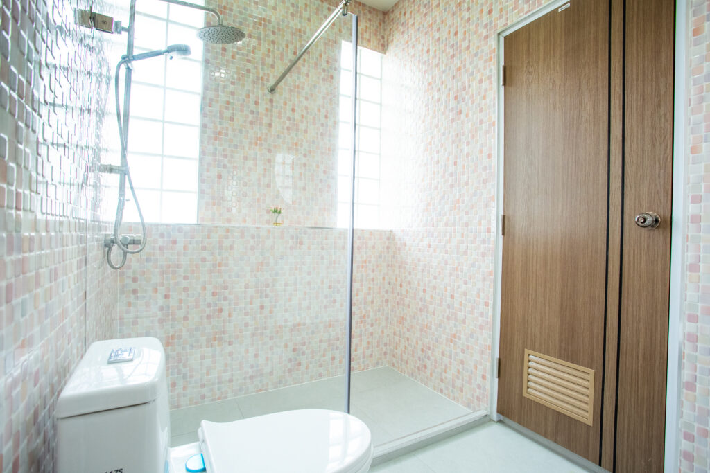 A modern, well-lit bathroom at Villa de L’Amitié Huahin featuring mosaic-style wall tiles, a glass shower partition, and a wooden door.