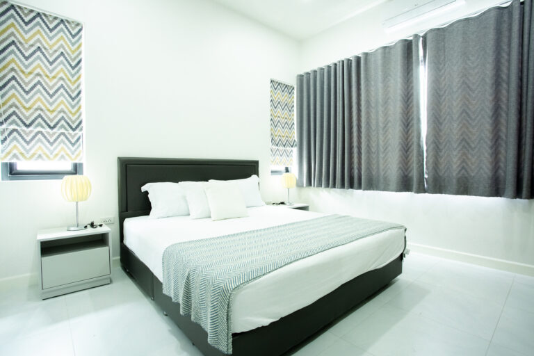 A modern minimalist bedroom at Villa de L’Amitié Huahin featuring a large bed with a light blue throw, grey curtains, and clean white walls.