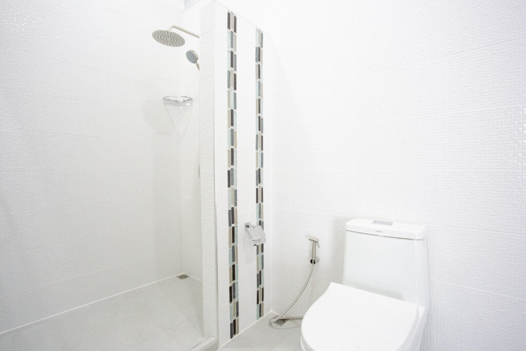 A bright and modern minimalist bathroom at Villa de L’Amitié Huahin featuring a glass-enclosed shower, neutral-toned decorative tile accents, and clean white fixtures.