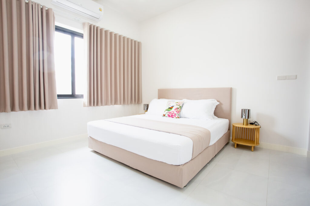 A bright, minimalist master bedroom at Villa de L’Amitié Huahin featuring a king-sized bed with neutral tones, wooden side tables, and elegant pleated curtains.