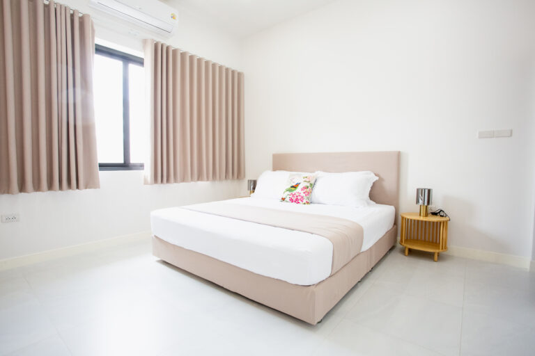 A bright, minimalist master bedroom at Villa de L’Amitié Huahin featuring a king-sized bed with neutral tones, wooden side tables, and elegant pleated curtains.