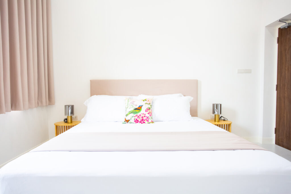 A cozy and bright bedroom at Villa de L’Amitié Huahin featuring a king-sized bed with white linens, modern bedside lamps, and warm neutral curtains.