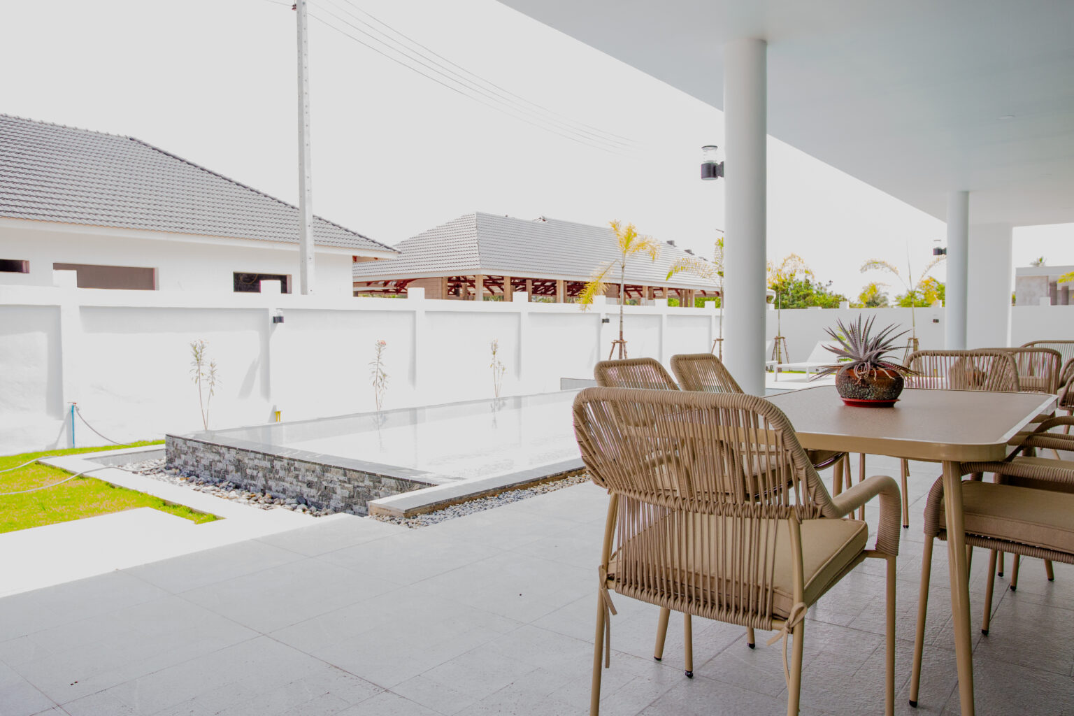 Close-up of a contemporary woven dining set on a tiled patio next to a minimalist private pool with stone waterfall feature at Villa de l'Amitie in Hua Hin, against a modern white boundary wall.